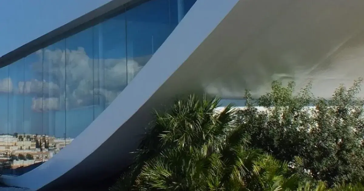 Arquitetura moderna, vidro reflete céu, palmeiras em primeiro plano.